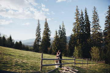 Wedding in the mountains. A young couple is standing near the fence against the backdrop of the forest and mountains at sunset and holding a bouquet in their hands