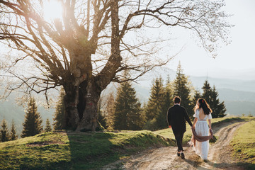 Wedding in the mountains. Young couple holding hands and running along a mountain road on the background of a large old tree