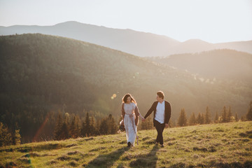 Fototapeta premium Wedding in the mountains. A guy and a girl in a white dress run along the road against the backdrop of mountains at sunset