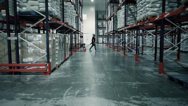 An operator walks with an electric pallet truck in a warehouse. The operator walks between products and pallets which are stocked in big shelving. Company and warehouse logistics