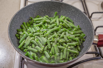 Fresh juicy green french beans are fried in a frying pan on a kitchen gas stove