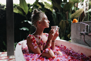 Refined girl sitting in bath on nature background. Wonderful caucasian lady relaxing during spa and drinking tea.