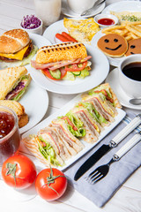 Breakfast on the dining table,. There are sandwiches, submarine sandwiches, burgers, pancakes, eggs, vegetables and coffee