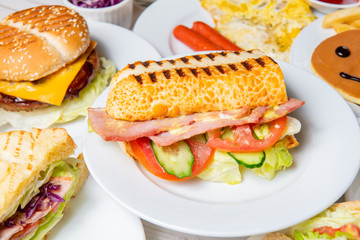 Breakfast on the dining table,. There are sandwiches, submarine sandwiches, burgers, pancakes, eggs, vegetables and coffee