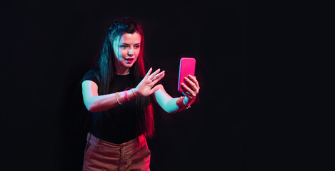 Fototapeta Tik tok advertisement concept. Young girl posing with smart phone in her hands, making selfie on black background. TIK TOK is a popular social network on the internet.  obraz