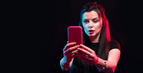 Tik tok advertisement concept. Young girl posing with smart phone in her hands, making selfie on black background. TIK TOK is a popular social network on the internet. 
