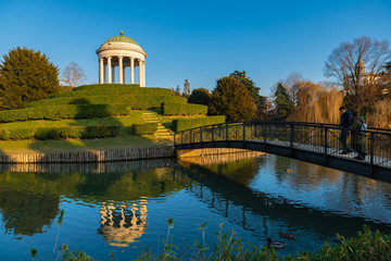 Fototapeta na wymiar A little monopteros ionic style temple is reflected on the lake of Parco Querini, the main public park of Vicenza city. A sunny and cloudless winter day. Couples of mallards swim quiet on water.