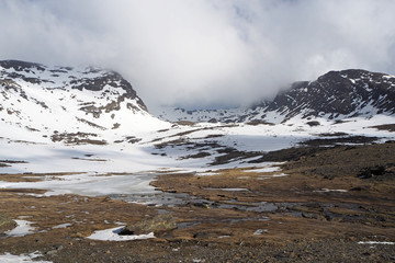 The mountain valley with the small river, dark rocky slopes covered by white snow, the blue sky with white clouds.