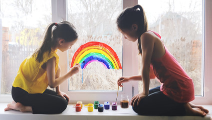 Two girls paint a rainbow on a window squatting