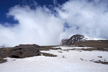 The mountain landscape with the dark rocks covered by white snow, the far peak, the blue sky with white clouds on the sunny day.