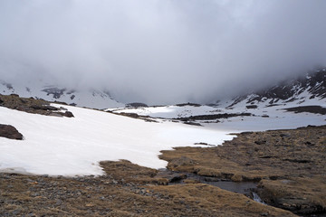 The mountain valley with the small river, the dry grass, the dark rocky slopes covered by white snow, the cloudy sky with low grey clouds.