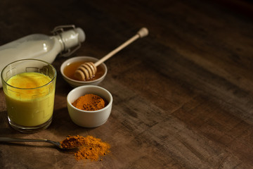 Turmeric Milk is poured into a glass. Next to it are the ingredients for making a drink - milk in a bottle on a napkin, spices and honey. Golden drink on dark moody background, selective focus. Top