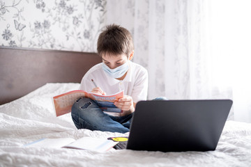 the boy sits at home on the bed in a sterile mask and undergoes online training on a laptop. Distance learning, online lessons. Quarantine and self-isolation during the school year