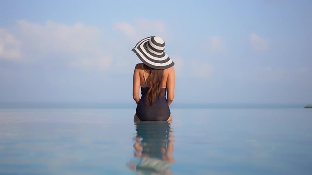 Woman sitting with back to camera on edge of infinity pool overlooking a scenic island beach view with blue sky and soft clouds. Water of swimming pool ripples in foreground lady tanning background.