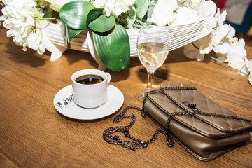A сup of coffee with lemon and sugar next to a glass of white wine, bouquet of artificial flowers and a woman's bag