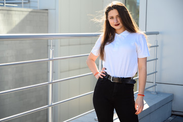 Fototapeta na wymiar Portrait of a caucasian brunette girl in a white t-shirt outdoors on a sunny day in the city against the background of the railing of the building.