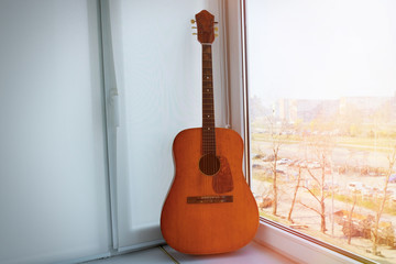 An old wooden, classic guitar standing by the window on a sunny day. Musical instrument.