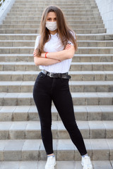 Portrait of a Caucasian young brunette girl in a mask from a virus in a white T-shirt outdoors in the afternoon in the city against the background of a building staircase.