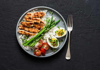 Balanced healthy lunch - rice, asparagus, grilled chicken, boiled egg on a dark background, top view