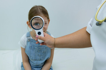 pediatrician examines in his office a little girl in denim overalls. Girl sitting on a couch in the white office of an ophthalmologist