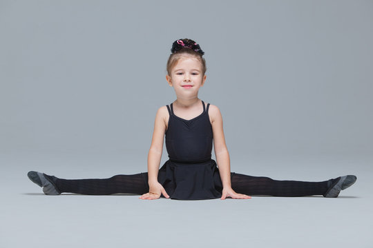 Beautiful little gymnast in black sportswear shows exercises on grey background. Flexible girl doing the splits to become ballet dancer