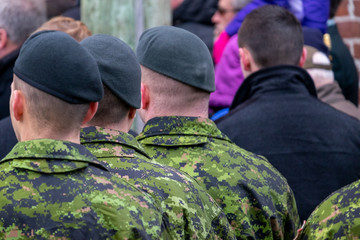 Fototapeta na wymiar Military soldiers wearing green and black camouflage uniforms in a crowd of people back on to the camera. The men are lined up shoulder to shoulder. The soldiers have dark black wool beret hats. 