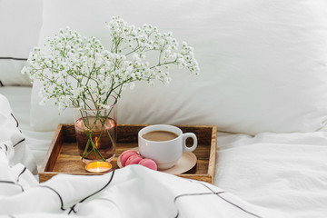 Wooden tray of coffee and candles with flowers on bed. White bedding sheets with striped blanket...