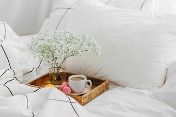 Wooden tray of coffee and candles with flowers on bed. White bedding sheets with striped blanket and pillow. Stay Home, quarantine.