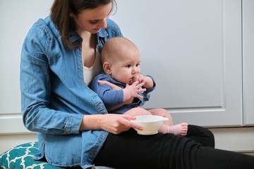 Young mother holding baby in arms, feeds toddler son with spoon