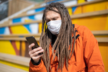 guy with dreadlocks in a white medical mask is reading news from a smartphone.