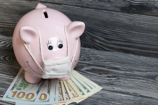 Ceramic piggy bank in pink. With a gauze bandage. In front of her are a few dollar bills. Savings during an epidemic. It stands on black painted boards.