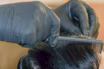 Hair dye color applied on the scalp by a hairdresser wearing black rubber gloves. At-home hair coloring styling, using a comb on the hair roots.