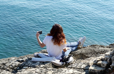 Young red haired woman taking a selfie on the beautiful stone beach near blue sea surface on the sunny day