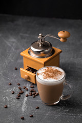 Side view of delicious cappuccino coffee with milk foam sprinkled with cinnamon in a transparent glass mug and wooden manual coffee grinder on a gray background, vertical format