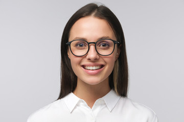 Headshot of young smiling business female, wearing glasses and white collar shirt, feeling confident, staying positive, isolated on gray background