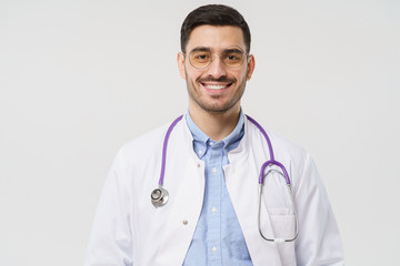 Fototapeta premium Close-up shot of handsome young male doctor with stethoscope around neck, wearing white coat and round glasses, isolated on gray background