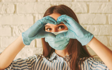 Happy girl in medical face mask and protective gloves looks through hands in shape of heart symbol....