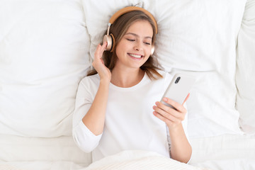 Girl with headphones lying in white bed after awaking, enjoying music that she chooses from smartphone via wireless connection, smiling happily in the morning