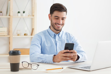 Business guy dressed in blue shirt sitting at desk in his office, holding phone and smiling happily while reading funny content on screen