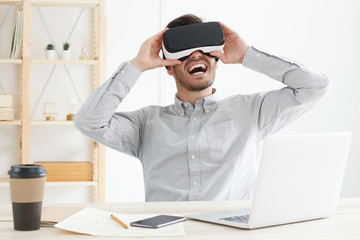 Young IT man laughing happily while exploring opportunities of virtual reality gaming with new headset on, spending free time in office