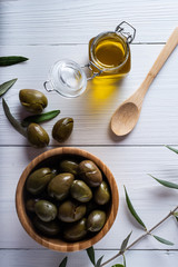 group of olives on a wooden vol next to a glass jar with oil on a white wooden table