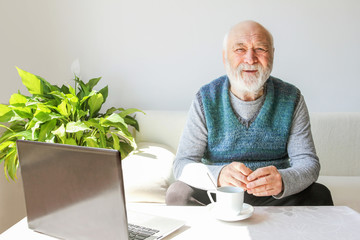 An elderly man while isolating a coronovirus, positively sits at home on a sofa in a bright interior, waiting for the end of the pandemic. Senior citizen 80 years in everyday life.
