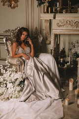 Magnificent young woman in luxurious dress and precious jewelery is sitting in a chair in a luxury apartment with lighted candles. Classic vintage interior. Beauty, fashion.