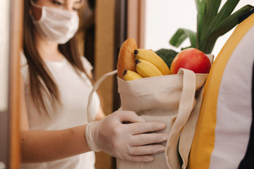 Delivery man in mask and gloves give fresh food to recipient and young woman customer. Stay home,...