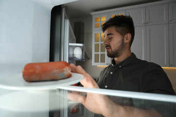 Young man feeling bad smell of spoiled sausage in refrigerator, view from inside - obrazy, fototapety, plakaty