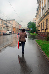 a woman with a bag and a red bag under a checkered umbrella walks in the rain down the street