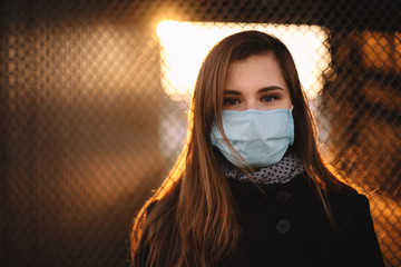 Portrait of young woman wearing protective face medical mask while standing against fence at sunset