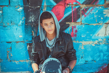 Portrait of a young handsome italian boy with skate and hood posing in the city urban graffiti...