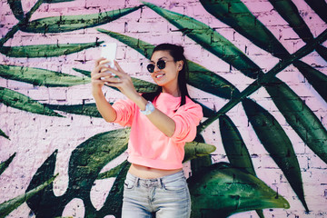 Carefree millennial content maker shooting influence video vlog near photo zone enjoying networking lifestyle, toothy Asian hipster girl in sunglasses smiling at smartphone camera for selfie image
