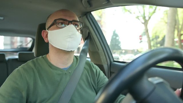 Turin, Piedmont, Italy. April 2020. Coronavirus pandemic: portrait of a Caucasian man driving the car wearing a white mask to avoid contagion. Selective focus on man and blurred background.
